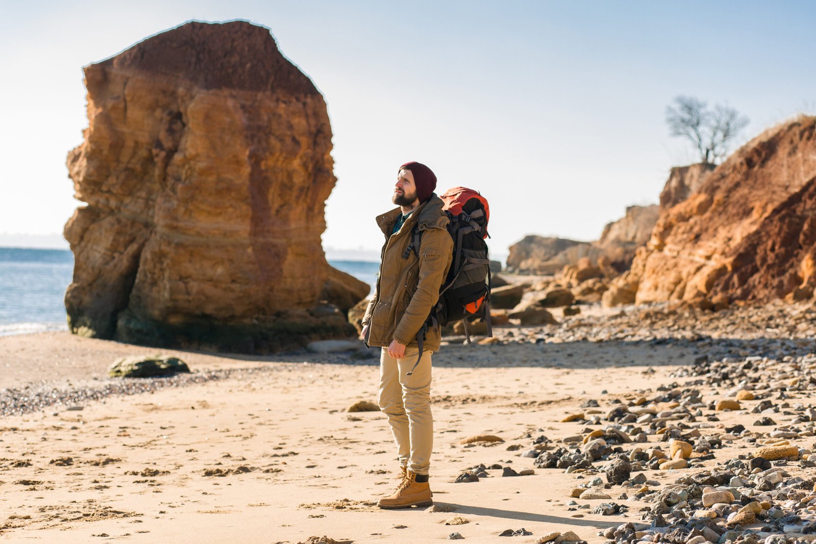 young hipster man traveling alone with backpack in autumn sea coast wearing warm jacket and hat, active tourist, exploring nature in cold season